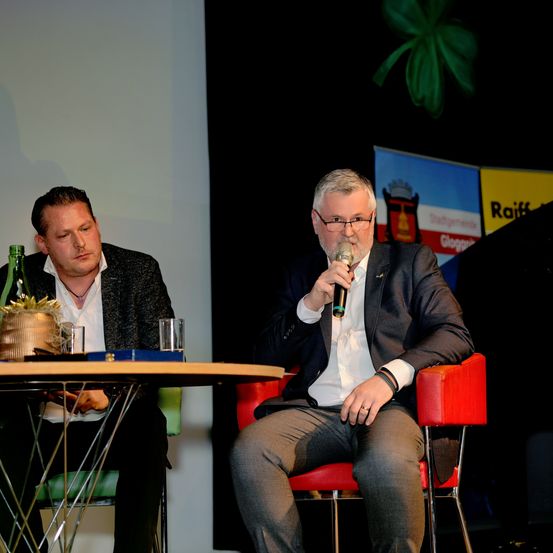 Zwei Männer sitzen in Stühlen an einem Tisch, einer mit einem Mikrofon und der andere mit einer Weinflasche und einem Topf. Dahinter ist eine Fahne mit einem Wappen und dem Wort Raiff an einer Wand angebracht.