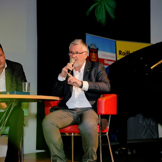 Ein Mann in einem Anzug sitzt auf einem roten Stuhl und hält ein Mikrofon, möglicherweise spricht er bei einer Veranstaltung. Ein anderer Mann sitzt neben ihm auf einem grünen Stuhl. Ein Tisch mit einem Glas steht vor ihnen. Dahinter befindet sich ein schwarzer Hintergrund mit Bannern.