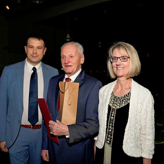Drei Menschen stehen in formeller Kleidung bei einer Veranstaltung zusammen; der Mann in der Mitte hält eine Urkunde und einen Beutel und lächelt. Die Frau neben ihm trägt eine Brille und eine weiße Jacke. Im Hintergrund ist eine dunkle Umgebung zu sehen.