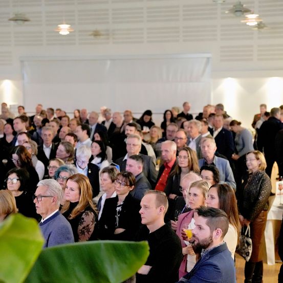 Eine große Gruppe von Menschen steht in einem Raum, vermutlich bei einer formellen Veranstaltung. Einige tragen Brillen und einige halten Getränke. Im Vordergrund befindet sich eine Pflanze.
