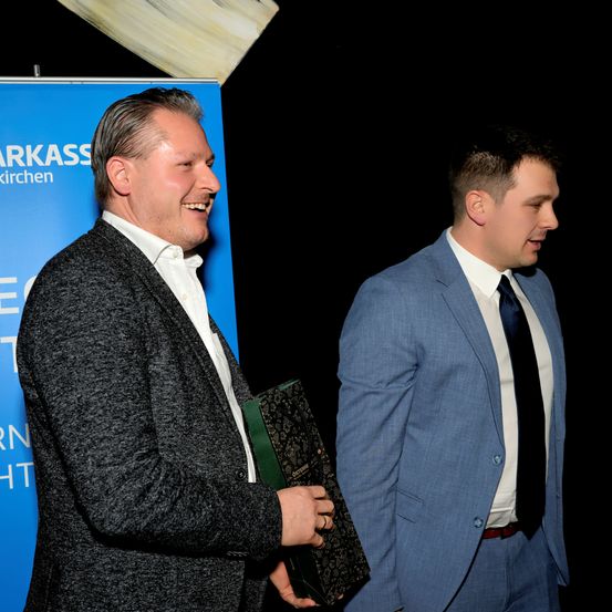 Zwei Männer in formeller Kleidung stehen nebeneinander. Einer der Männer hält eine Mappe, und beide lächeln. Sie scheinen sich auf einer Veranstaltung zu befinden. Ein blauer Banner mit weißem Text befindet sich hinter ihnen.
