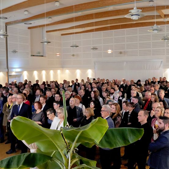Eine große Menschenmenge, darunter Männer und Frauen, steht drinnen, einige halten Gläser. Eine grüne Pflanze ist im Vordergrund. Die Decke ist hölzern, mit hängenden Lampen.