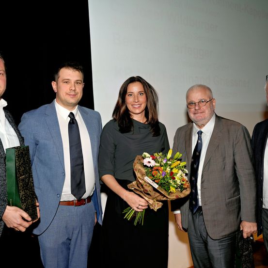 Eine Gruppe von fünf Personen steht vor einer Projektionsleinwand. Die Frau in der Mitte hält einen Blumenstrauß. Der Mann auf der linken Seite hält eine grüne Mappe. Der Mann auf der rechten Seite trägt eine Brille.