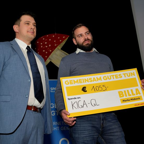 Zwei Männer stehen zusammen, einer hält einen großen gelben Scheck in der Hand, auf dem 'Gemeinsam Gutes Tun' steht und ein Spendenbetrag von 1.055 Euro vermerkt ist. Der Mann im Anzug lächelt, während der bärtige Mann ernst aussieht. Hinter ihnen befindet sich ein Hintergrund mit einem Pilz.