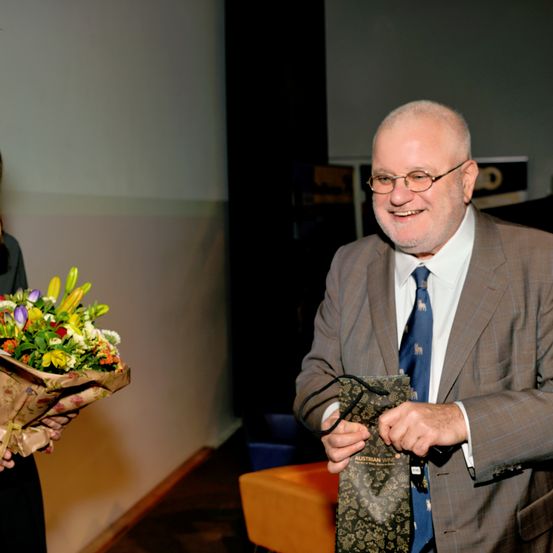 Ein älterer Mann mit Brille hält eine dekorative Weintasche und lächelt, während er einen Blumenstrauß von einer anderen Person erhält.
