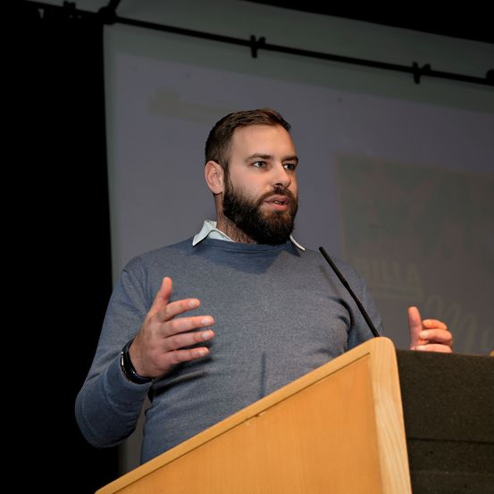 Ein Mann mit Bart steht an einem Rednerpult und spricht in ein Mikrofon. Er gestikuliert mit den Händen. Das Pult ist aus Holz und eine Projektionsleinwand ist hinter ihm zu sehen.