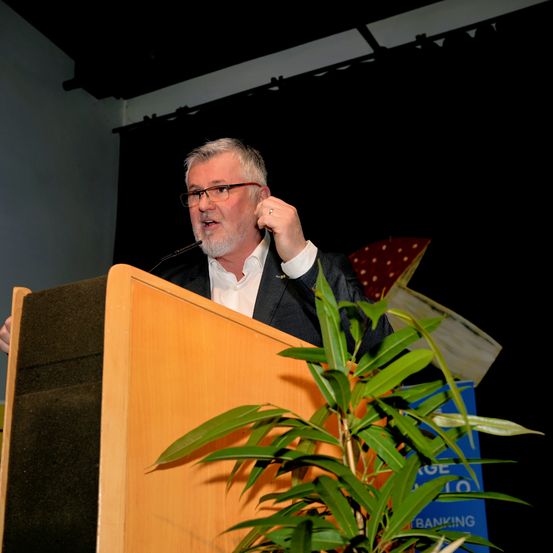 Ein Mann mit Brille und weißem Hemd steht hinter einem Podium mit Mikrofon. Eine Pflanze mit grünen Blättern steht vor dem Podium. Dahinter befindet sich eine Wand mit einem schwarzen Vorhang.