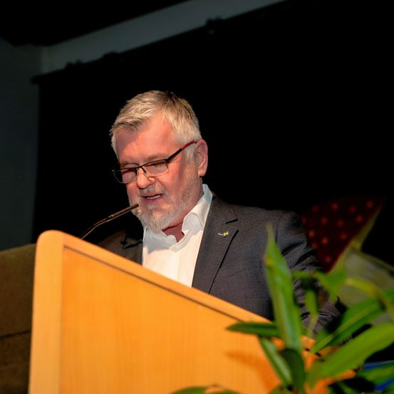 Ein Mann mit Brille und Anzug spricht an einem Rednerpult mit Mikrofon. Er lächelt.