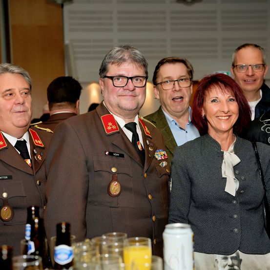 Fünf Erwachsene stehen bei einem formellen Ereignis um einen Tisch. Der Mann in Uniform lächelt, während die Frau neben ihm zufrieden aussieht. Sie sind von Gläsern, einer Dose und Flaschen mit Getränken umgeben. Ein weiterer Mann hinter ihnen scheint zu sprechen.