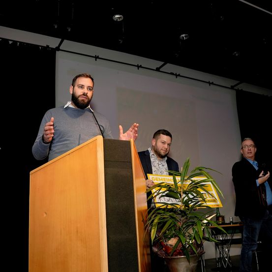 Ein Mann steht an einem Rednerpult und spricht vor einem Publikum. Hinter ihm halten zwei andere Männer ein Schild mit der Aufschrift 'Gemeinde'. Vor ihnen steht eine Topfpflanze. Die Umgebung ist ein Raum mit einer Projektionsleinwand und schwarzen Vorhängen.