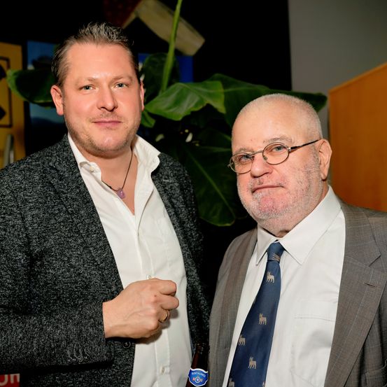 Zwei Männer stehen nebeneinander, einer hält eine Bierflasche, während der andere eine Brille trägt und eine Krawatte mit Hirschmuster. Sie lächeln und scheinen für ein Foto zu posieren. Hinter ihnen steht eine große Pflanze und eine Holzstruktur.