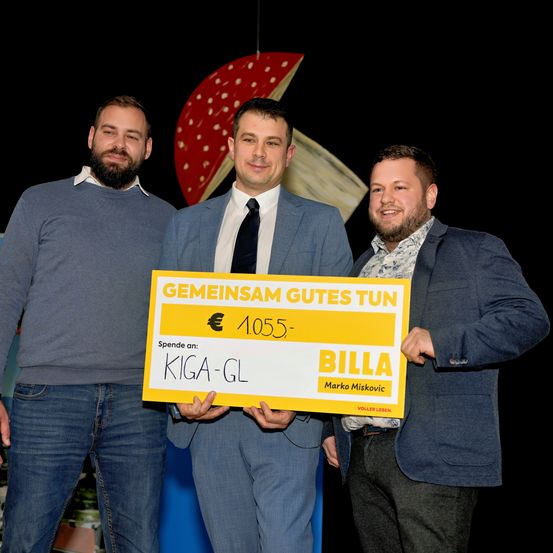 Drei Männer stehen zusammen und halten einen großen Scheck über 1.055 Euro. Der Mann in der Mitte hält den Scheck hoch und lächelt. Der Mann auf der linken Seite hat einen Bart und trägt ein graues Hemd. Der Mann auf der rechten Seite trägt einen blauen Anzug.