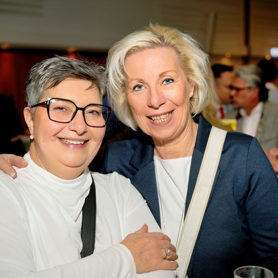 Zwei Frauen lächeln und posieren für ein Foto, eine mit Brille und weißem Shirt, die andere in einem blauen Blazer. Sie sind bei einer Veranstaltung mit Menschen hinter ihnen.