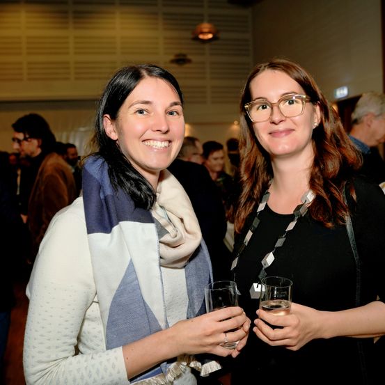 Zwei Frauen in einem überfüllten Raum, sie lächeln und halten Gläser mit Wasser in der Hand. Die Frau auf der linken Seite trägt einen weißen und blauen Schal. Hinter ihnen stehen und unterhalten sich Menschen.