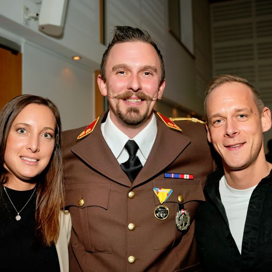 Ein Mann in Uniform posiert mit einer lächelnden Frau und einem anderen Mann bei einer Veranstaltung.