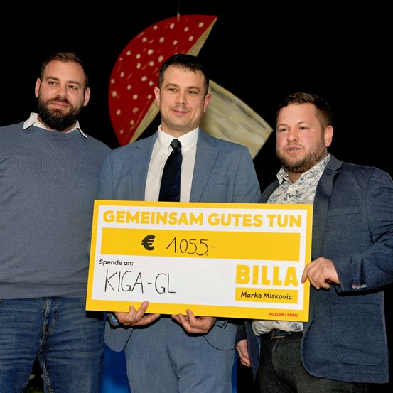 Drei Männer halten ein gelbes Zertifikat mit den Worten 'GEMEINSAM GUTES TUN' und 1.055 €. Sie lächeln und schauen in die Kamera. Dahinter befindet sich ein rot-goldenes Objekt.