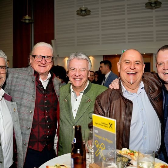 Fünf Männer lächeln und posieren für ein Foto bei einer Veranstaltung, zwei von ihnen tragen Brillen. Einer hält eine Bierflasche, und andere haben Teller mit Essen. Ein Schild mit Logo und Text steht auf dem Tisch.