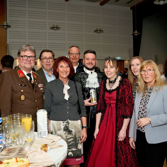 Eine Gruppe Erwachsener posiert für ein Foto bei einer Veranstaltung, einige in formeller Kleidung. Ein Tisch davor hat Getränke und Essen.