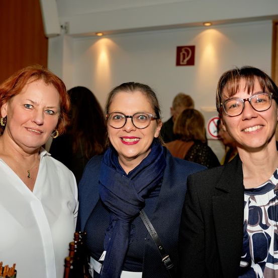 Drei Frauen stehen nebeneinander und lächeln für die Kamera. Die Frau auf der linken Seite trägt ein weißes Hemd und Ohrringe. Die Frau in der Mitte trägt einen blauen Schal und eine Brille. Die Frau auf der rechten Seite trägt einen schwarzen Mantel und eine Brille. Hinter ihnen stehen weitere Personen.