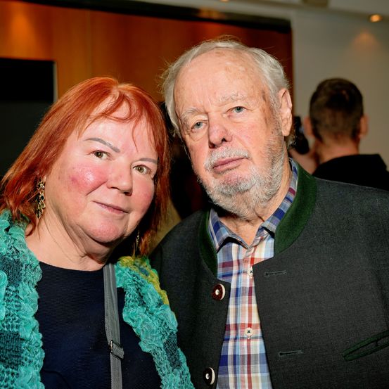 Ein älterer Mann und eine Frau stehen bei einer Veranstaltung eng beieinander und lächeln. Der Mann hat einen Bart und trägt ein kariertes Hemd und eine schwarze Jacke. Die Frau hat rote Haare und trägt einen grünen Pullover. Hinter ihnen macht ein Mann ein Foto.