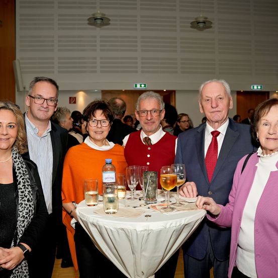 Eine Gruppe von Menschen, darunter zwei Frauen und drei Männer, stehen um einen Tisch herum mit Weingläsern, Flaschen und anderen Gegenständen. Sie sind feierlich gekleidet, wobei zwei Männer Krawatten tragen.