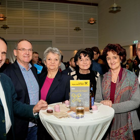 Fünf Menschen, vier Frauen und ein Mann, stehen gemeinsam in einem Raum. Sie lächeln und scheinen sich bei einer Veranstaltung zu befinden. Ein Tisch vor ihnen hat Tassen, ein Glas und eine gelbe Box mit Text. Hinter ihnen sind andere Menschen im Hintergrund.