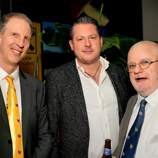 Drei Männer in Anzügen, die zusammenstehen. Der linke trägt eine gelbe Krawatte, der mittlere einen grauen Mantel und der rechte eine Brille. Sie lächeln, möglicherweise für ein Foto. Hinter ihnen ist eine Pflanze und ein gelbes Schild zu sehen.