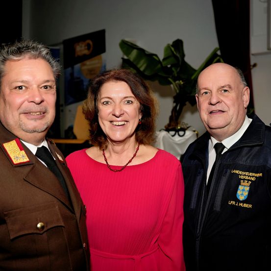 Drei Personen lächeln und posieren für ein Foto. Eine Frau in einem roten Kleid steht in der Mitte, mit einem Mann in einer braunen Uniform auf ihrer linken Seite und einem Mann in einem schwarzen Jackett auf ihrer rechten Seite. Sie stehen vor einer Pflanze und einer Wand mit einem Schild.