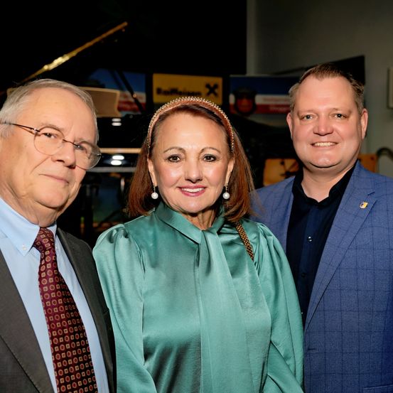 Drei Personen stehen lächelnd zusammen, wahrscheinlich bei einer Veranstaltung. Ein Mann links trägt eine Brille und eine gemusterte Krawatte. Die Frau in der Mitte hat eine Stirnband und trägt ein grünes Kleid. Der Mann rechts trägt einen blauen Anzug. Ein Klavier und ein Schild sind hinter ihnen.