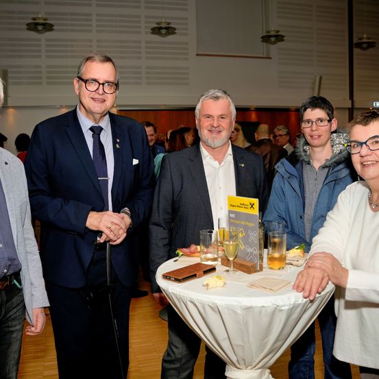 Eine Gruppe von Menschen bei einer Veranstaltung. Zwei Männer in Anzügen stehen hinter einem Tisch mit Getränken. Ein Mann mit Brille schaut in die Kamera.