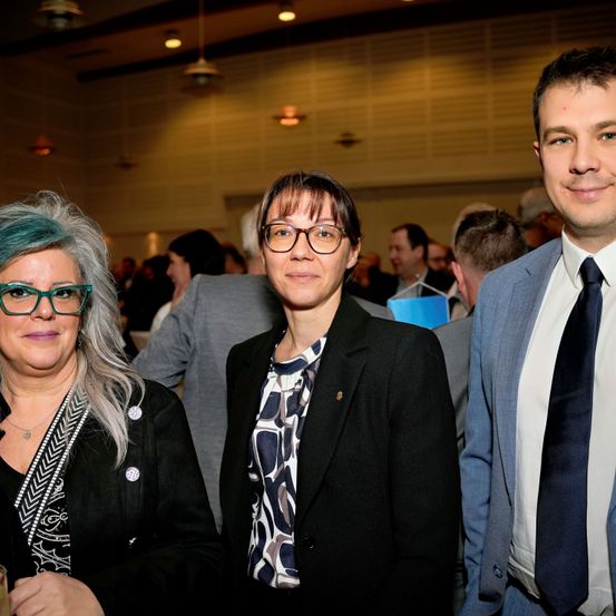 Drei Menschen stehen bei einer formellen Veranstaltung zusammen. Die Frau auf der linken Seite hat blaue Haare und trägt eine Brille. Die Frau in der Mitte hat eine Brille und ein gemustertes Oberteil. Der Mann auf der rechten Seite lächelt und trägt einen Anzug mit Krawatte.