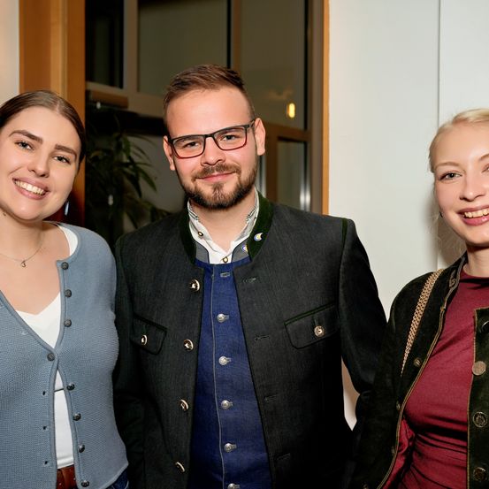 Drei Personen lächeln für ein Foto, ein Mann in der Mitte und zwei Frauen an seinen Seiten. Der Mann trägt eine Brille und eine schwarze Jacke. Beide Frauen tragen Halsketten und Jacken, eine mit einem hellblauen Oberteil und die andere mit einem roten Oberteil. Hinter ihnen befindet sich ein Glasfenster.