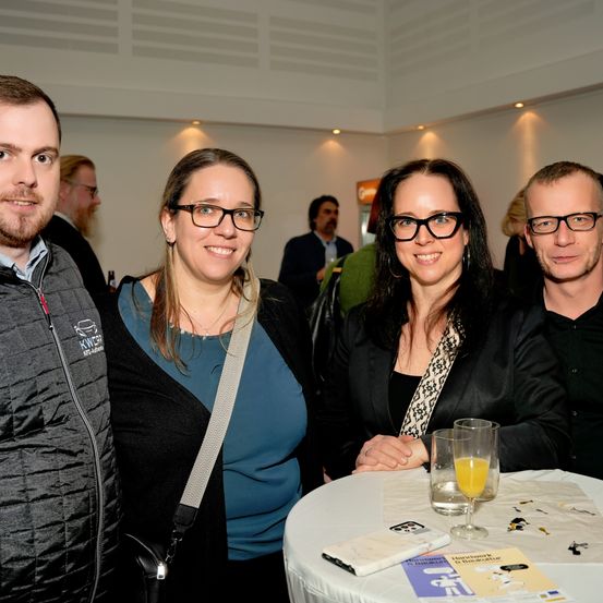 Vier Menschen lächeln für ein Foto an einem Tisch. Ein Mann und eine Frau links und eine weitere Frau und ein Mann rechts. Sie sind bei einer Veranstaltung mit einem unscharfen Hintergrund.