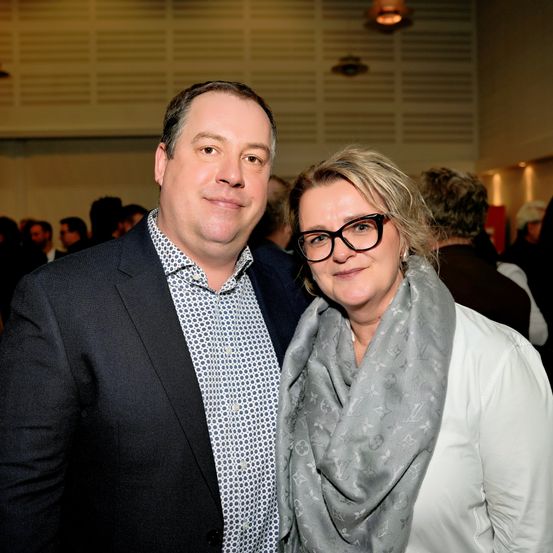 Ein Mann und eine Frau stehen bei einer Veranstaltung eng beieinander und lächeln für die Kamera. Der Mann trägt einen schwarzen Anzug und die Frau ein weißes Hemd und einen grauen Schal. Sie sind von anderen Menschen umgeben.