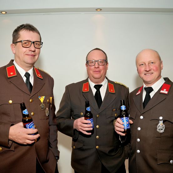 Drei Männer in Militäruniformen halten Bierflaschen in der Hand und stehen nebeneinander. Sie alle lächeln.