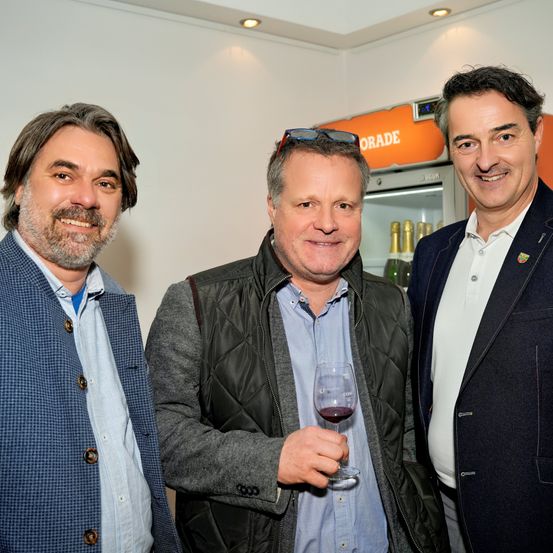 Drei Männer stehen in einem Raum und lächeln für ein Foto. Ein Mann hält ein Glas Rotwein. Hinter ihnen steht ein Kühlschrank mit einem Markennamen.