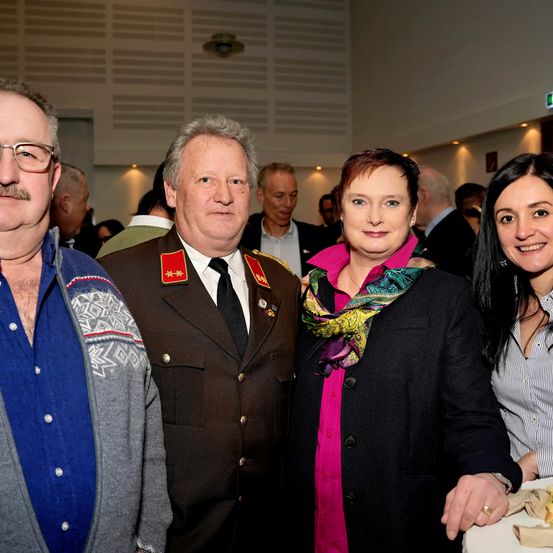 Vier Personen stehen eng beieinander in einer Innenumgebung. Ein Mann in Uniform, eine Frau in einem rosa Shirt und Schal und zwei weitere Frauen. Sie scheinen an einer Veranstaltung teilzunehmen.