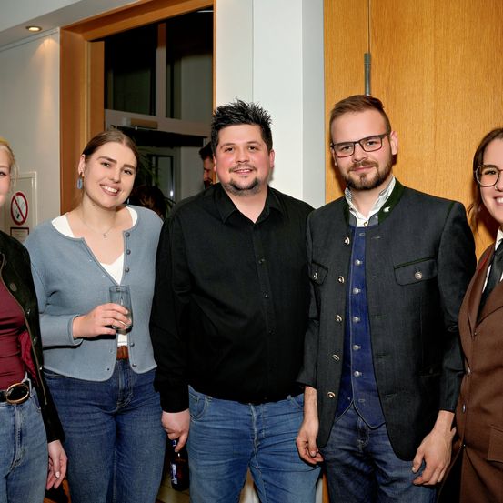 Fünf Menschen stehen lächelnd in einem Raum. Sie sind lässig gekleidet, eine Frau hält ein Glas. Hinter ihnen steht eine Holztür mit einer weißen Wand und einem Rauchverbotsschild.