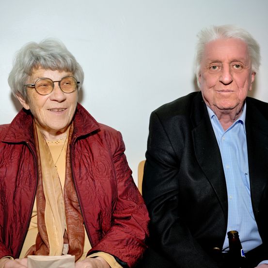 Ein älterer Mann und eine ältere Frau sitzen eng beieinander und lächeln. Die Frau trägt eine Brille und eine rote Jacke. Der Mann trägt einen schwarzen Anzug und ein blaues Hemd und hält eine Flasche. Sie sitzen vor einem schlichten weißen Hintergrund.