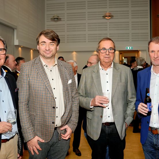 Vier Männer stehen eng beieinander und lächeln für ein Foto bei einer Veranstaltung. Der Mann auf der linken Seite hält ein Weinglas. Der zentrale Mann hat eine Flasche in der Hand, während der Mann auf der rechten Seite eine Flasche hält. Der Mann in der Mitte trägt eine Brille und einen Anzug.