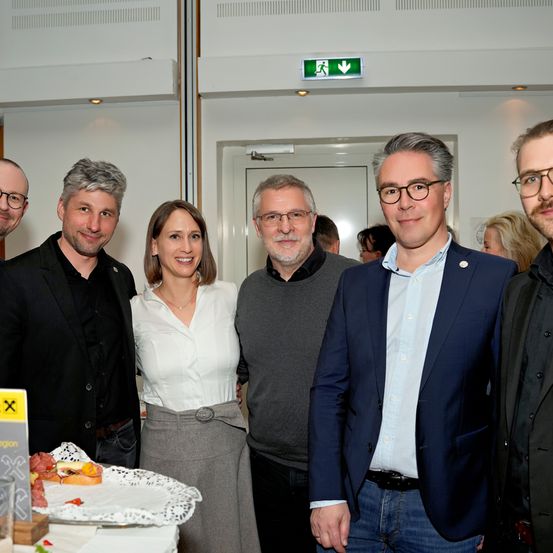 Fünf Menschen stehen lächelnd zusammen in einem Raum. Vor ihnen steht ein Tisch mit Essen. Sie tragen formelle Kleidung. Ein Ausgangsschild befindet sich an der Wand.