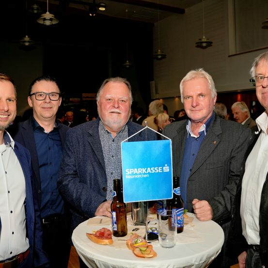 Fünf Männer stehen um einen Tisch mit Essen und Getränken und lächeln für ein Foto. Ein blaues Schild für Sparkasse Neunkirchen ist auf dem Tisch.