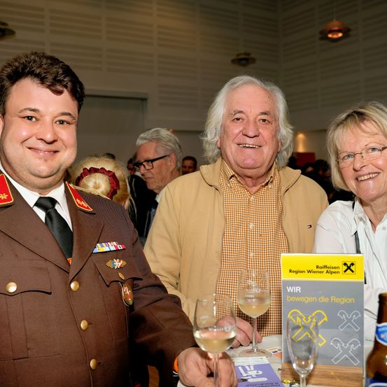 Drei Personen, darunter ein Militäroffizier, ein Mann in einem karierten Hemd und eine Frau, lächeln in die Kamera. Sie haben Gläser mit Wein auf dem Tisch.