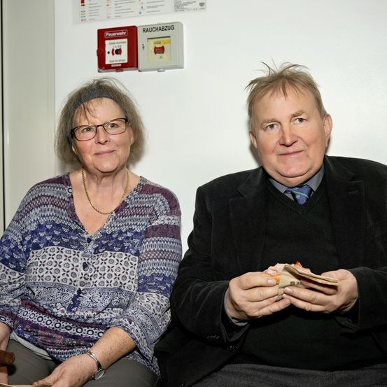 Ein älteres Paar sitzt auf einem Stuhl an einer weißen Wand. Ein Feuerlöscher ist über ihnen montiert. Die Frau trägt eine Brille und eine Halskette. Der Mann hält Geld in den Händen.