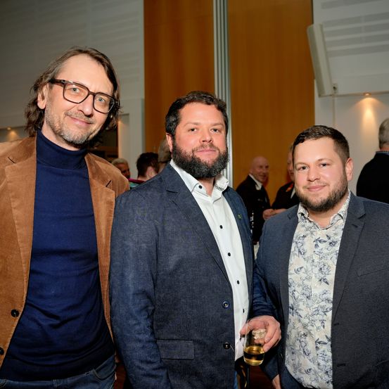 Drei Männer bei einer formellen Veranstaltung, zwei in Anzügen und einer in Rollkragenpullover und Jacke, lächelnd und eng beieinanderstehend, wobei der mittlere Mann ein Glas mit Getränk in der Hand hält.