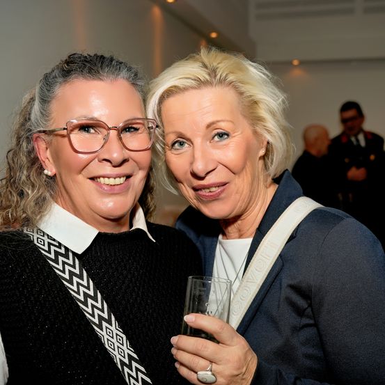 Zwei Frauen lächeln und posieren für ein Foto. Die Frau auf der linken Seite trägt eine Brille und eine gemusterte Umhängetasche. Die Frau auf der rechten Seite hält ein Glas und trägt einen Blazer. Hinter ihnen stehen Menschen in einem Raum.
