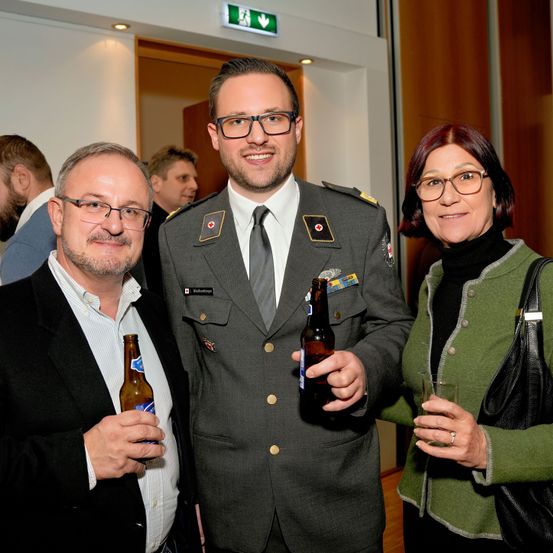 Drei Menschen stehen zusammen und lächeln. Ein Mann in Uniform und zwei weitere, einer mit einer Bierflasche, der andere mit einem Glas. Sie scheinen bei einer Veranstaltung zu sein.