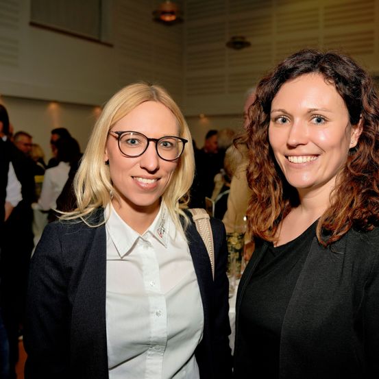 Zwei Frauen, die lächeln und sich bei einer Veranstaltung nahe stehen. Die Frau links trägt eine Brille und ein weißes Hemd. Die Frau rechts hat lockiges Haar und trägt ein schwarzes Oberteil. Im Hintergrund unterhalten sich mehrere Personen.