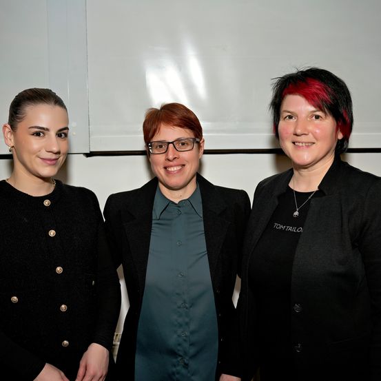Drei Frauen stehen nebeneinander, lächeln und posieren für ein Foto. Die Frau auf der linken Seite trägt einen schwarzen Cardigan mit goldenen Knöpfen. Die mittlere Frau trägt eine Brille und ein türkisfarbenes Hemd. Die rechte Frau hat rote Haare und trägt einen schwarzen Blazer.