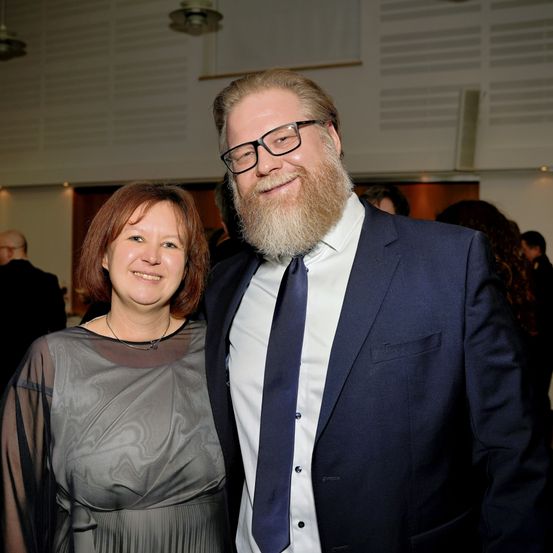 Ein Mann mit Bart und eine Frau in einem grauen Kleid lächeln und posieren für ein Foto. Sie stehen vor einer weißen Wand.